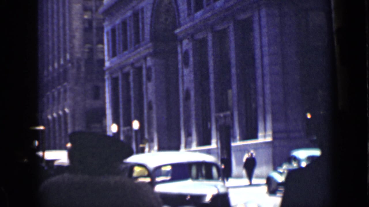 Shadowed pedestrians and 1930s cars along a city street beside tall masonry buildings.