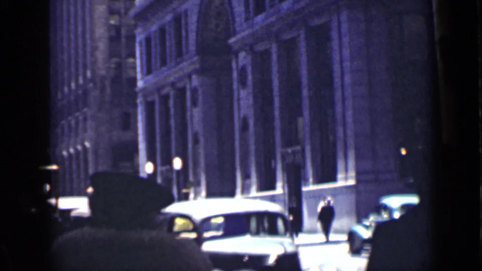 1939 San Francisco street view with pedestrians in hats, parked or passing automobiles, lit street lamps, and tall dark masonry buildings.