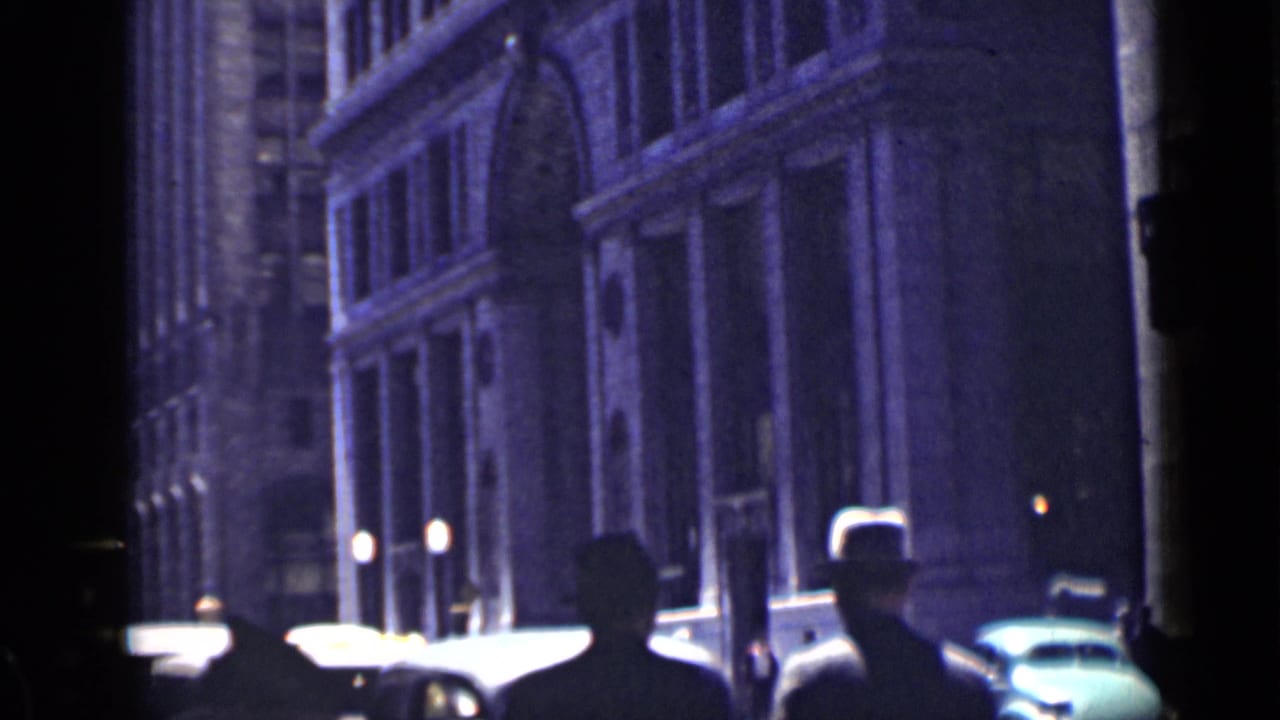 People in hats pass along a dim city street with cars and tall building facades.