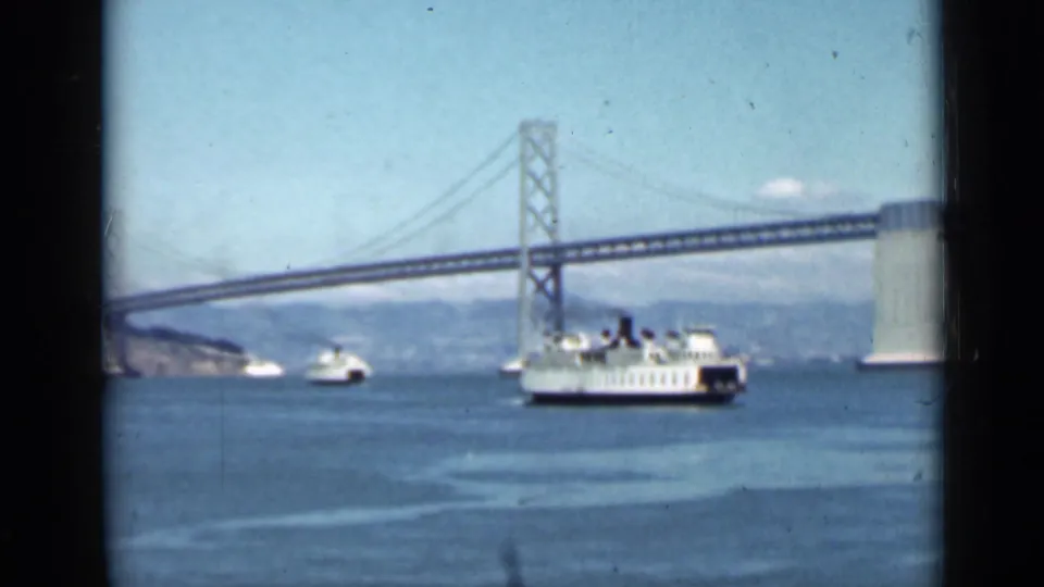 Wide 1939 San Francisco Bay view with the Bay Bridge, ferry traffic, open water, distant shoreline, and pale sky.