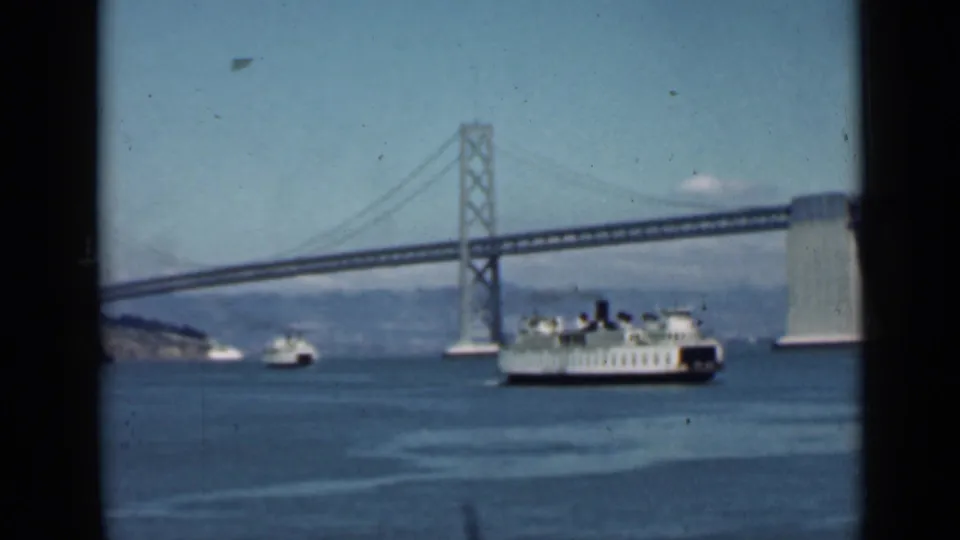 1939 San Francisco Bay view with a white ferry passing in front of the Bay Bridge tower and distant shoreline.