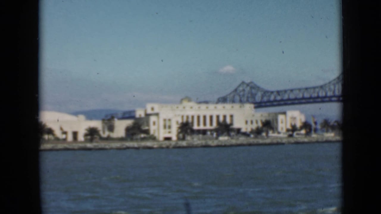 White waterfront buildings across blue water with a steel bridge span behind them.