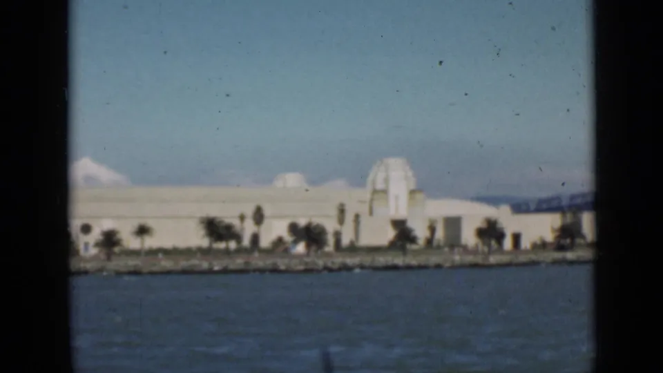 San Francisco waterfront from the bay, with a long white building, shoreline trees, and soft 8mm color.
