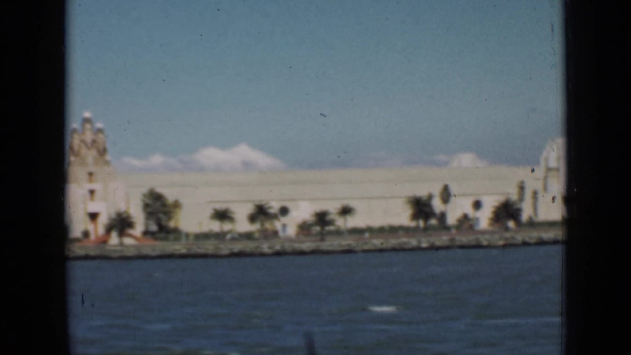 Waterfront building with tower detail, palm trees, bay water, and distant pale mountains.