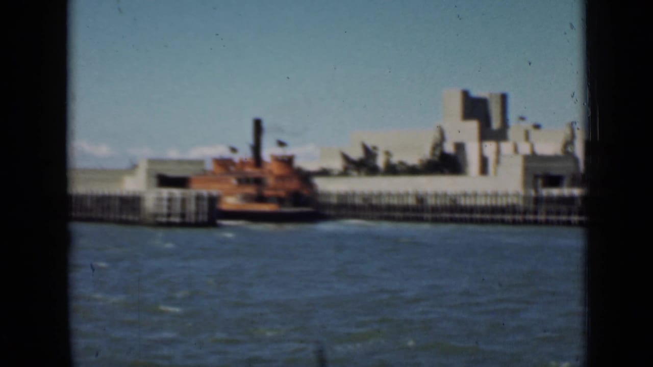 Red-brown boat near a pier with pale waterfront buildings behind it.