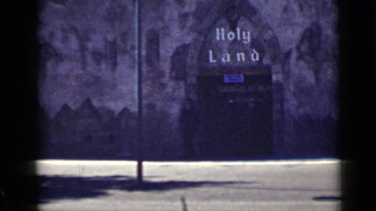Holy Land sign above a dark arched entrance beside a pale sidewalk edge.