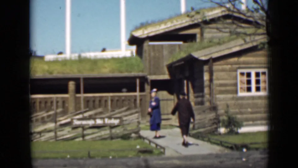 1939 San Francisco frame showing two women in period clothing near Norway’s Ski Lodge, a rustic wood building with a grass-covered roof.