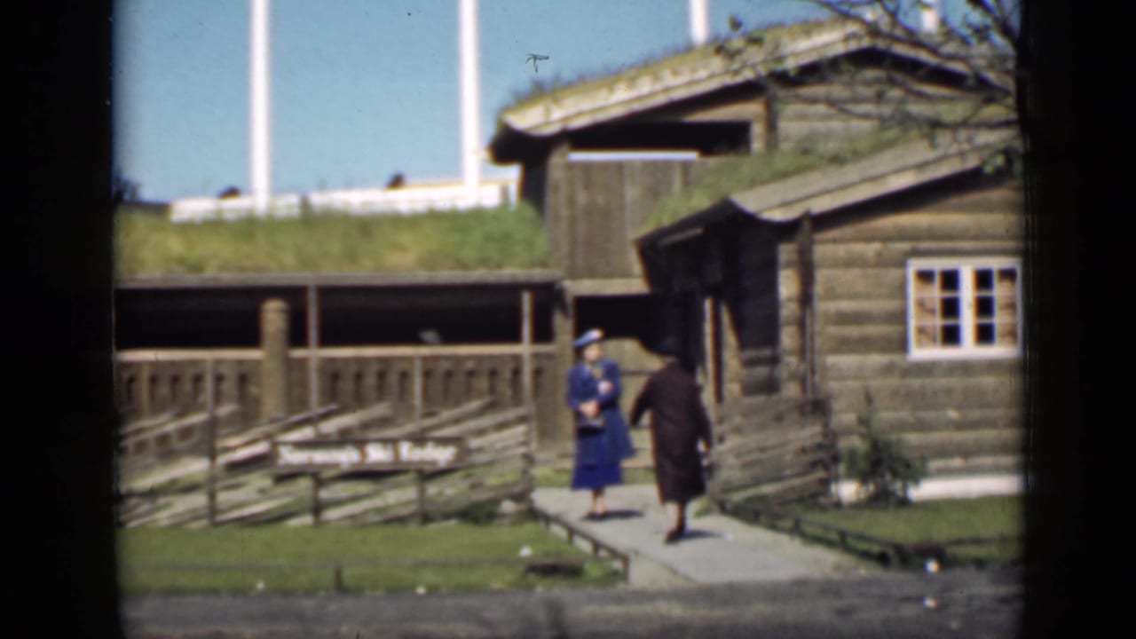 Two women walk on a path beside a rustic log building with a grass roof.