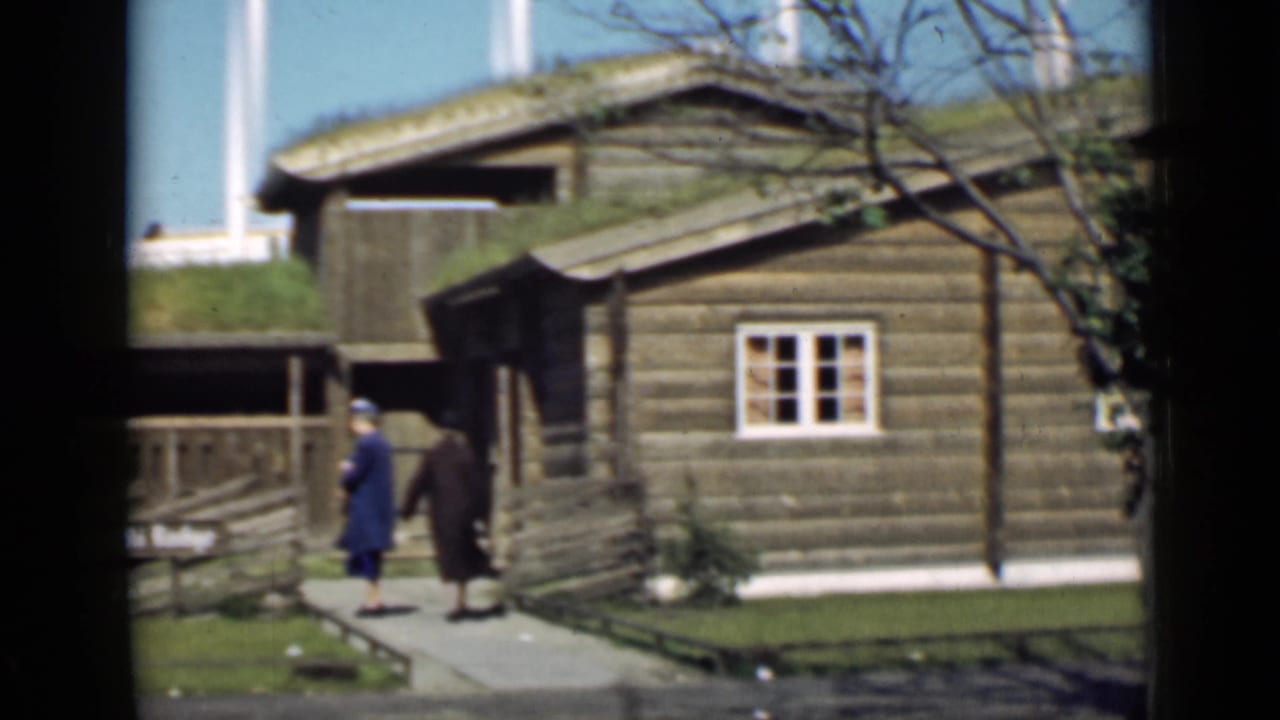 Two women stand or walk near the corner of a log building with a grass roof.