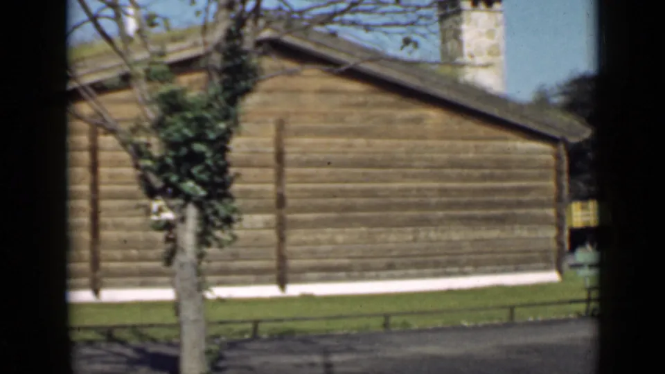 1939 San Francisco frame showing a long log wall, ivy-covered tree, stone chimney, lawn, and an unreadable yellow sign near the edge.