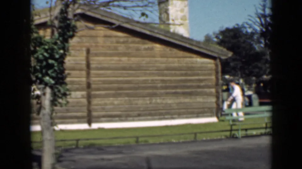 1939 San Francisco home-movie frame showing a man near a rustic wooden cabin, stone chimney, lawn, and green bench.