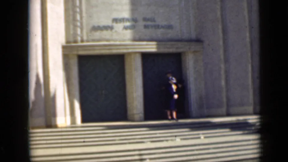 1939 San Francisco home-movie frame showing a woman in dark dress near festival hall doors and broad exterior steps.
