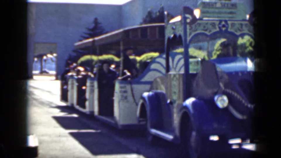 A close 8mm view shows the ornate sightseeing tram passing by, with riders seated along the open side and hedges behind it.