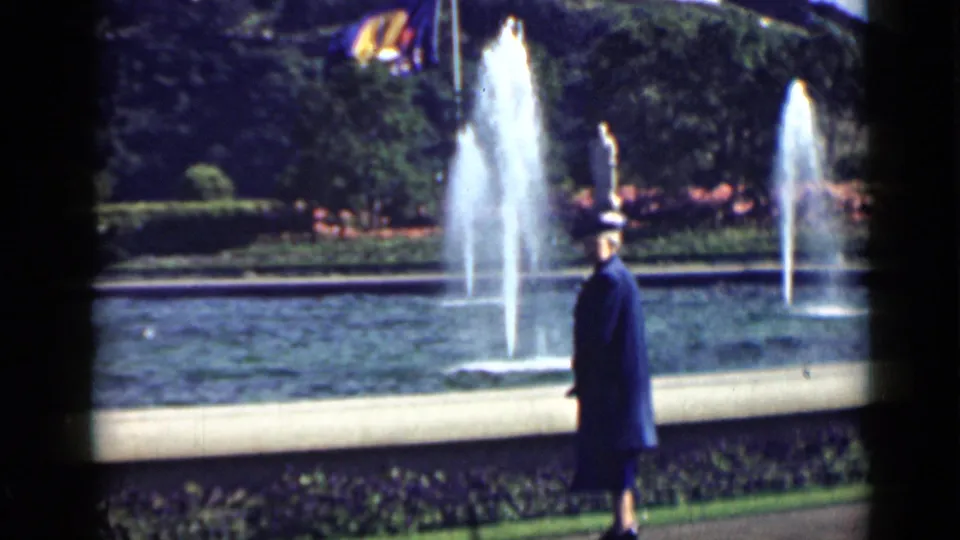 A woman in a dark coat strolls beside a landscaped fountain, with water jets, trees, and flowers visible in the background.