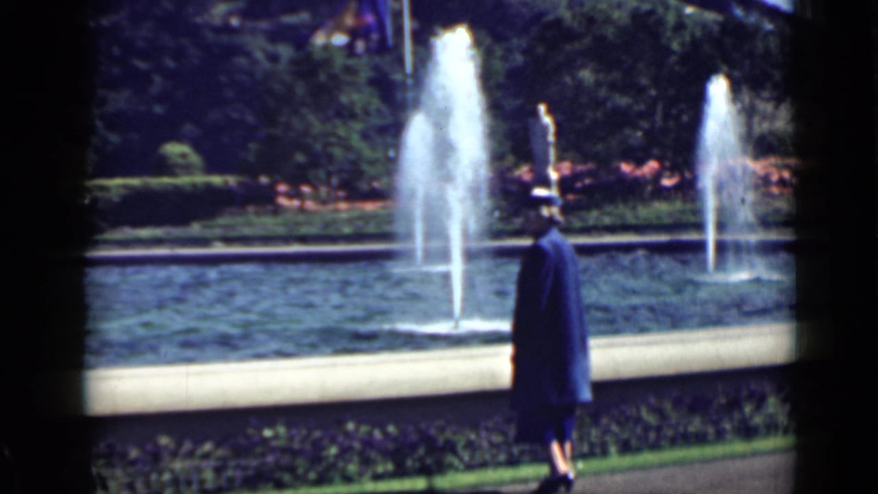Woman in a blue dress and hat stands beside a fountain pool with water jets and hedges behind her.