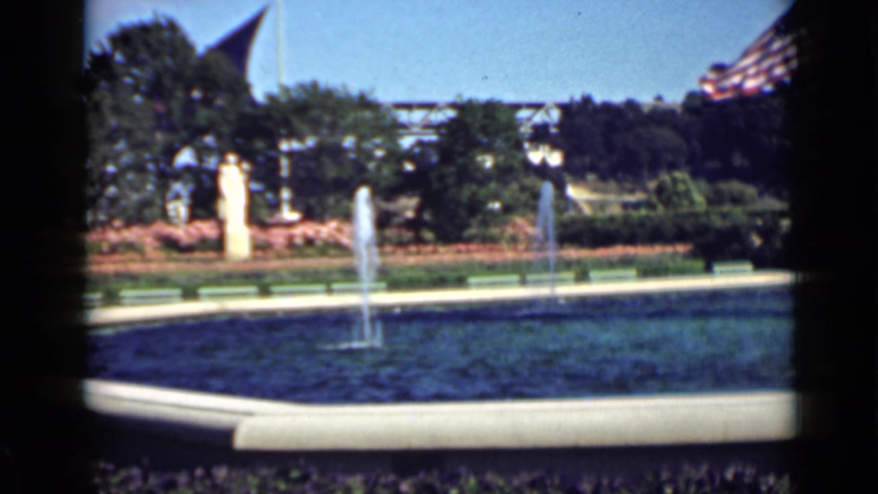 Fountain pool with water jets, shrubs, benches, trees, and American flags in a sunny park.
