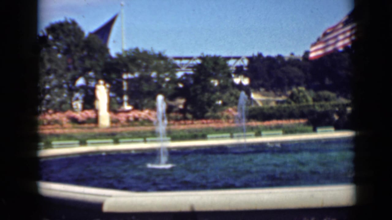 Park fountain with a central jet, shrubs, benches, trees, and a large American flag.