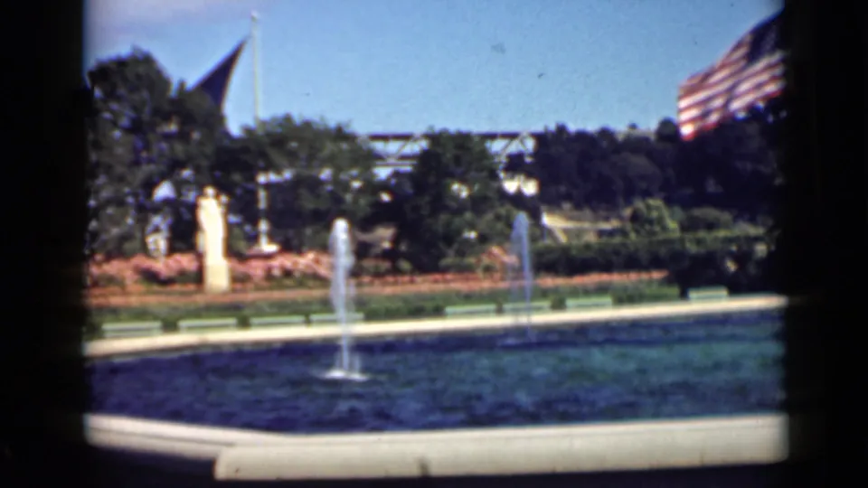 1939 San Francisco home-movie frame of a landscaped park fountain, low benches, flower beds or shrubs, trees, and American flags.