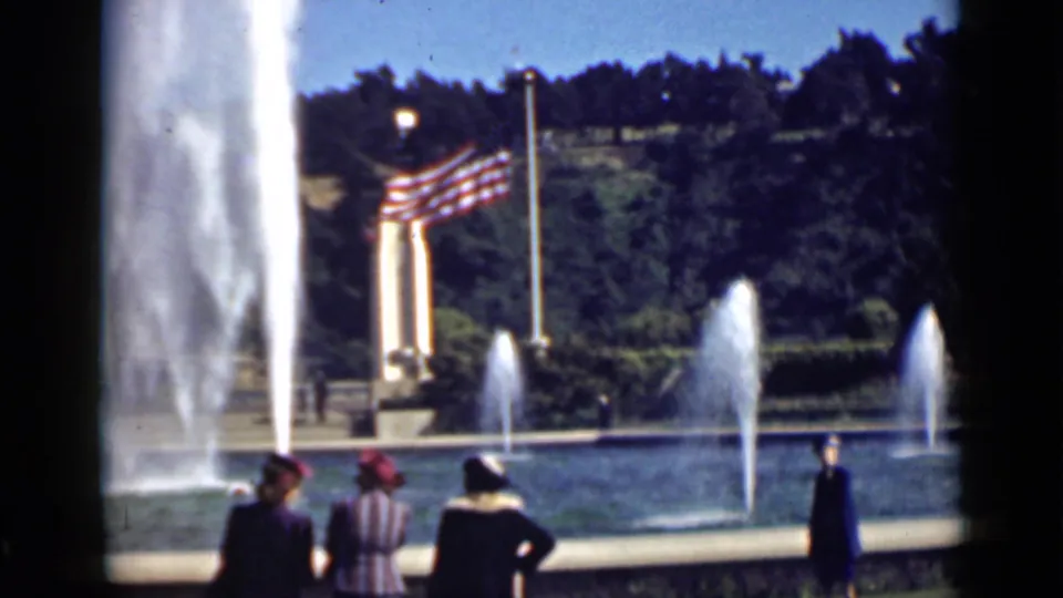 1939 San Francisco park visitors near a fountain, with tall water spray, American flags, trees, benches, and a partially legible sign.