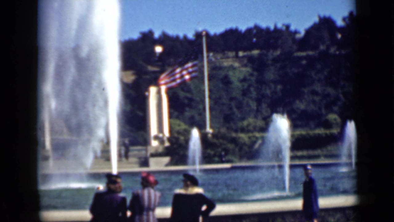 Visitors stand near a large fountain with an American flag and trees behind it.