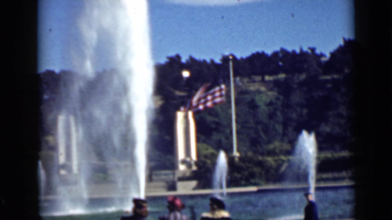 A fountain sprays beside an American flag while visitors pass along the pool edge.