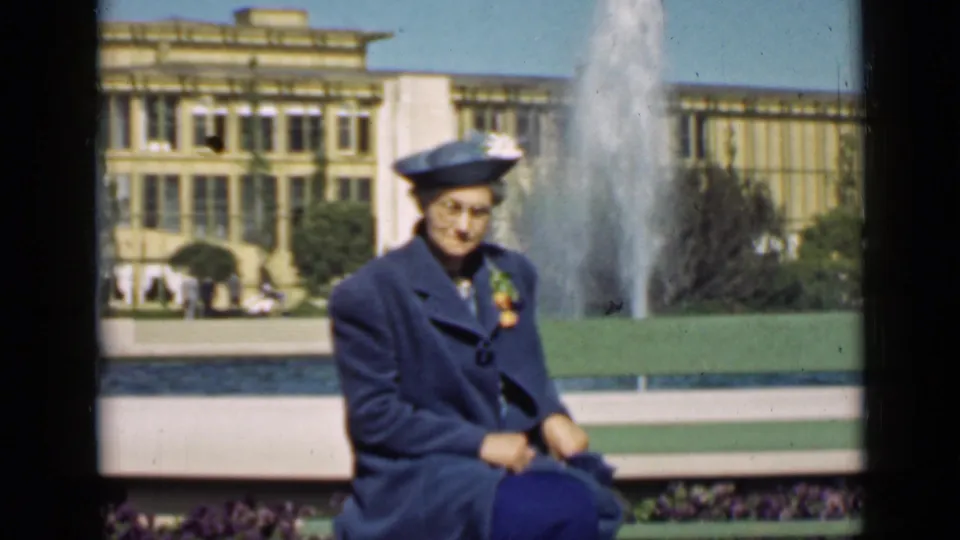 1939 San Francisco home-movie frame of a seated woman in a blue coat and hat near a fountain and large public building.