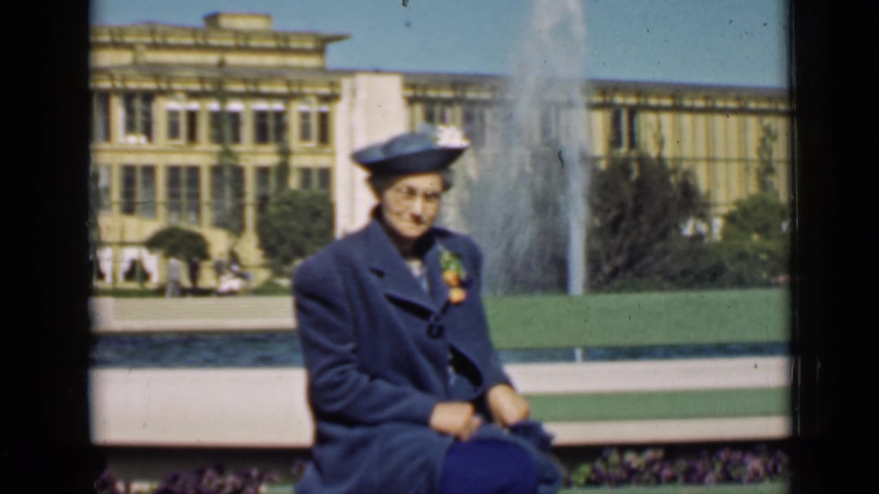 Woman in a blue coat and hat seated near a fountain and fairground building.