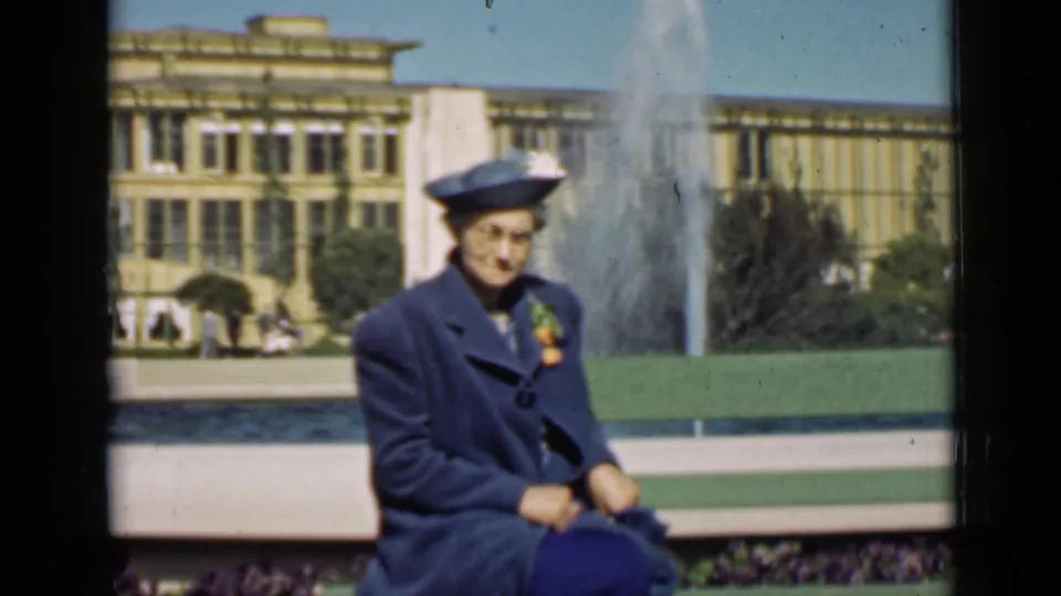Well-dressed woman in blue seated on a green bench near a fountain at the 1939 San Francisco World's Fair.