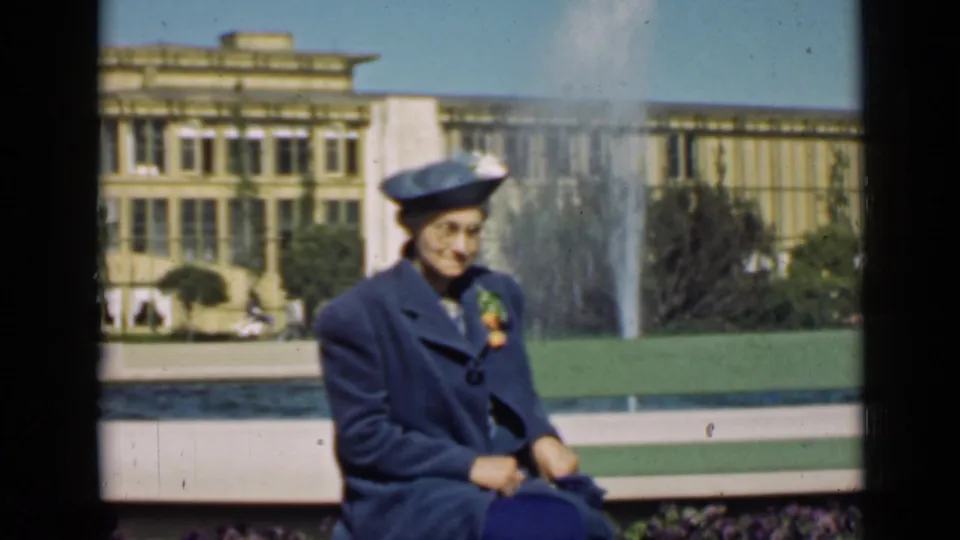 A woman in a blue coat and hat sits in front of a fountain with fairground architecture in the background.