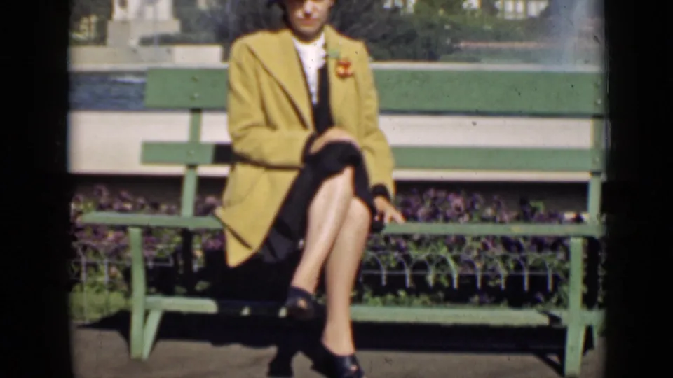 A well-dressed woman in a yellow coat sits cross-legged on a bench at the 1939 San Francisco World's Fair setting.