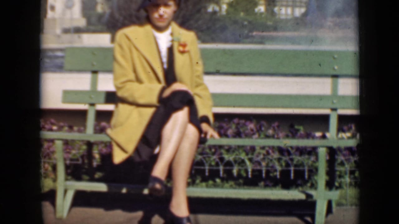 Woman in a yellow coat and dark hat sitting on a green bench near plantings.