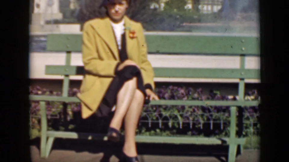 In 1939 San Francisco footage, a woman in a mustard-yellow coat sits posed on a green park bench with flowers behind her.