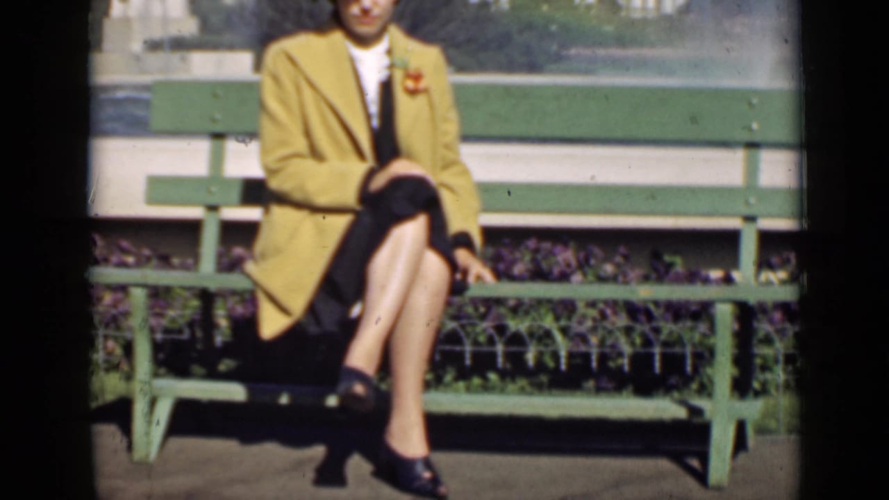 Woman seated on a green bench, wearing a yellow coat, dark skirt, and hat.