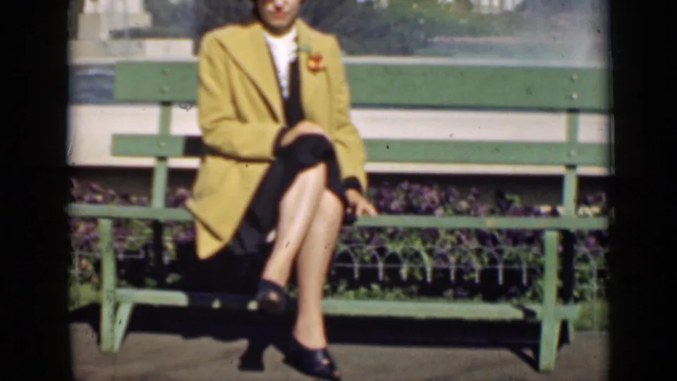 The woman remains seated on the green bench in San Francisco, her yellow coat and pinned flower visible in soft 8mm color.
