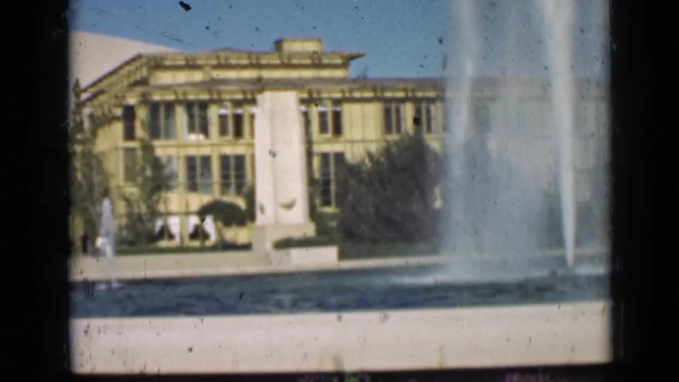 A fountain sprays across the foreground, with a large classical building and a few distant figures visible beyond it.