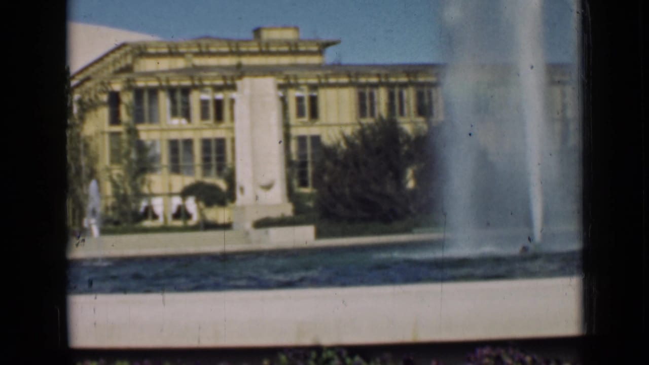 Tall fountain plume in front of a large pale public building and landscaped grounds.
