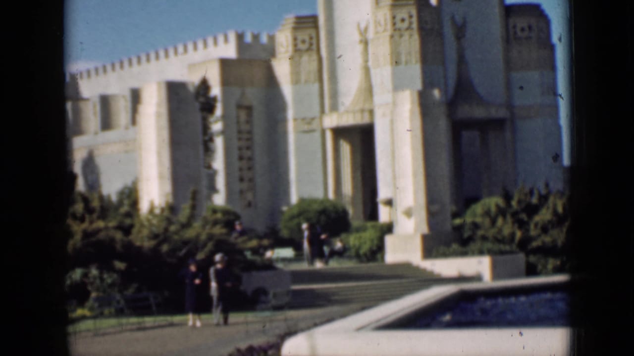 Ornate pale building entrance with columns, steps, shrubs, and visitors near a pool.