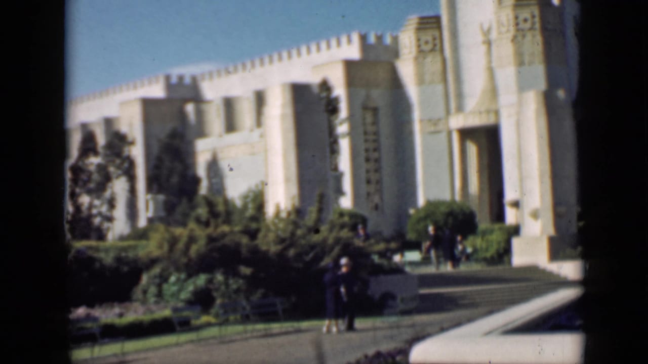Pale monumental building behind hedges and lawn with visitors on a paved path.