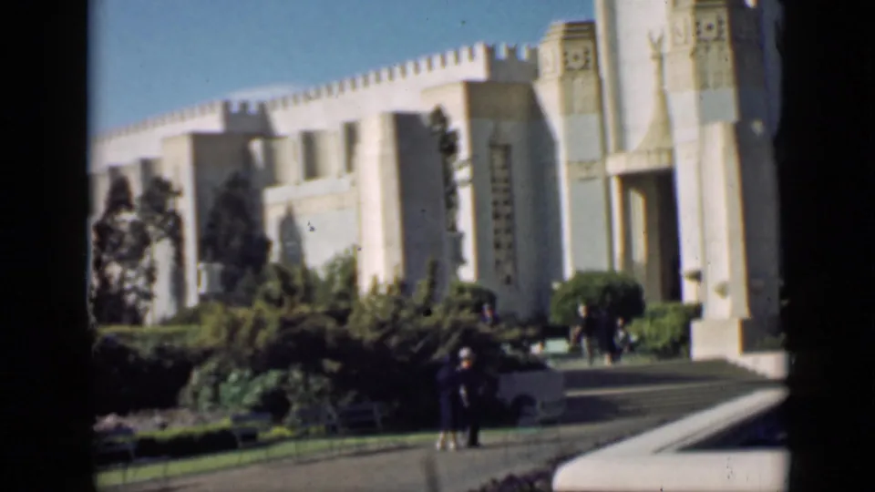 1939 San Francisco home-movie frame showing a pale monumental building, manicured greenery, benches, and distant visitors on a path.