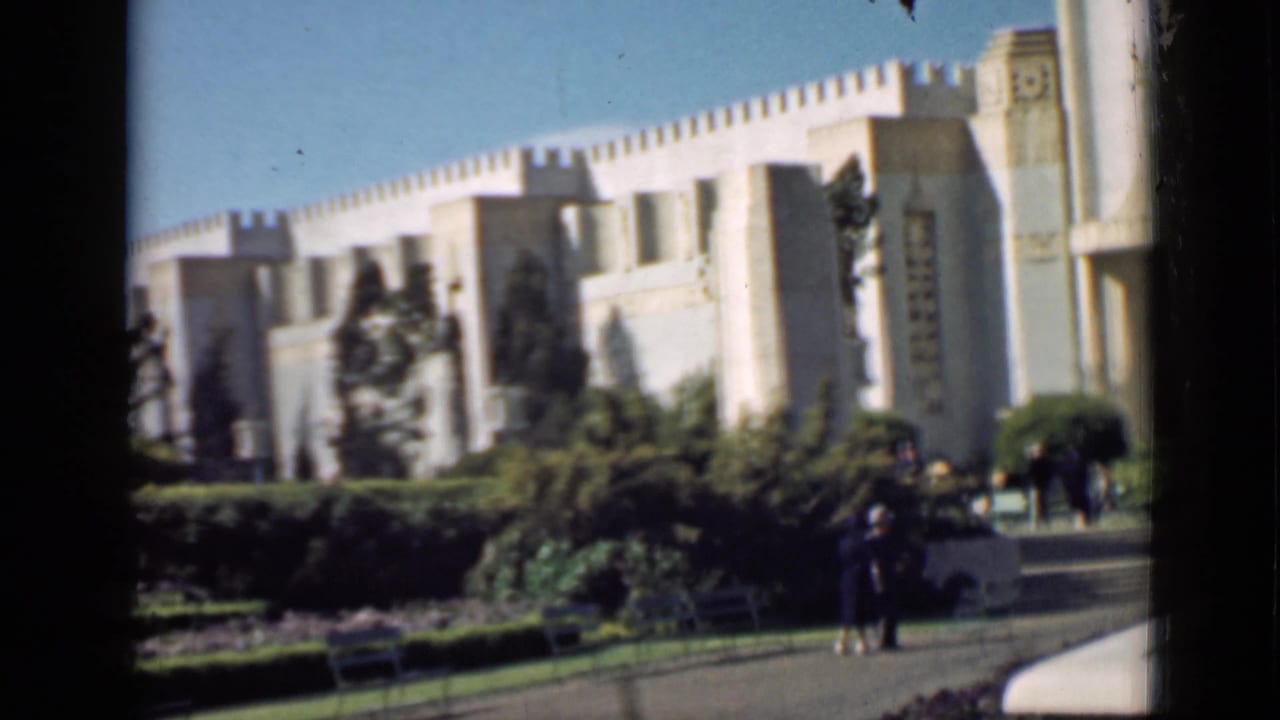 Large pale building with hedges, trees, lawn, and two visitors near the path.