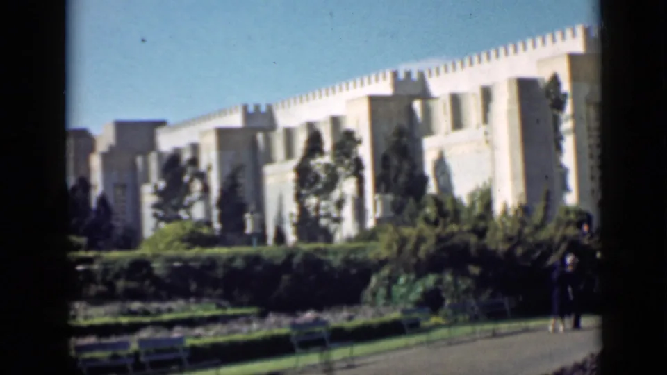1939 San Francisco home-movie view of a monumental pale facade seen across lawns, hedges, benches, and a distant visitor at the edge of the frame.