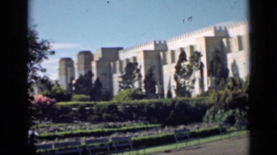 1939 San Francisco frame showing benches and manicured plantings in front of a pale monumental building under a clear blue sky.