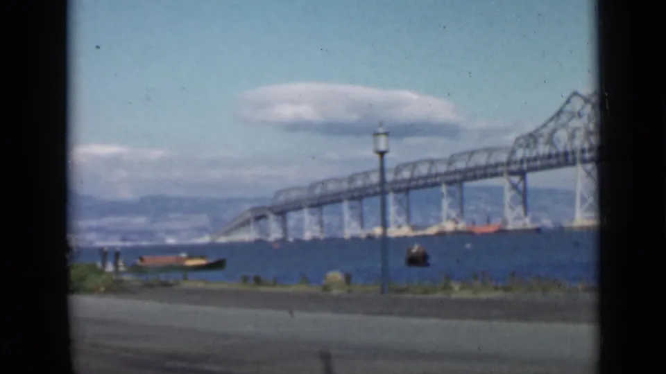 Wide 1939 San Francisco Bay view with a steel truss bridge, small boats, road edge, lamppost, and distant hills.