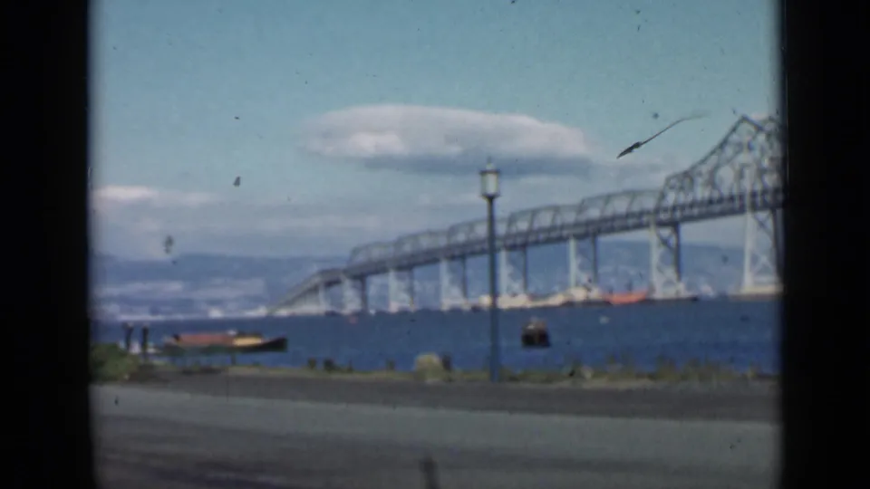 1939 San Francisco Bay frame showing the bridge structure, waterfront road, small boats, lamppost, clouds, and distant hills.