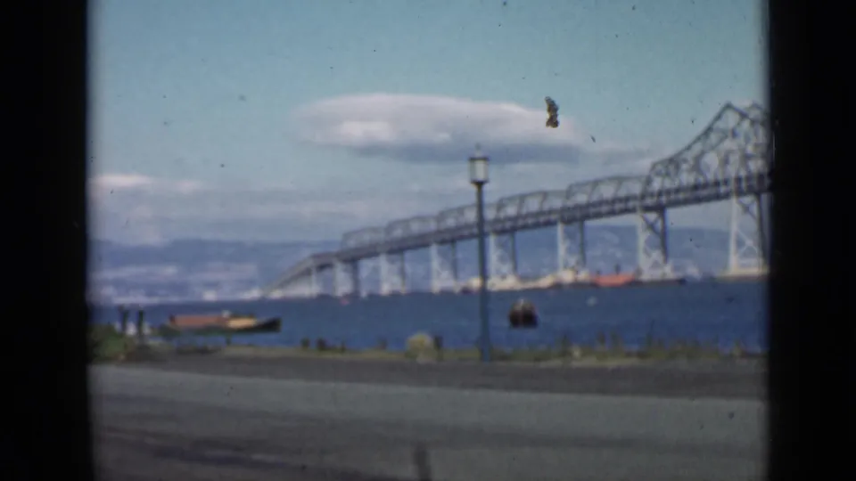 Soft 1939 San Francisco Bay home-movie frame with bridge trusses, boats on the water, a lamppost, road edge, and clouds.