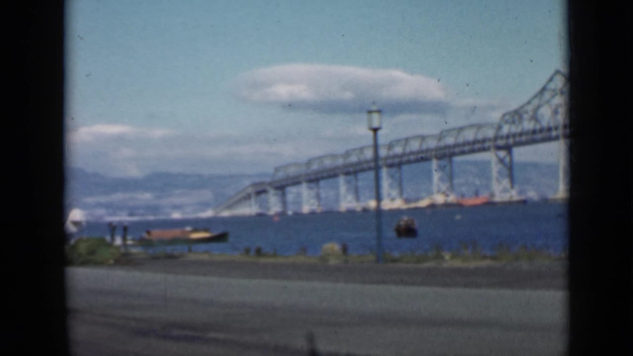 Large bridge over blue water with a seaplane, small boat, road, lamppost, hills, and clouds.