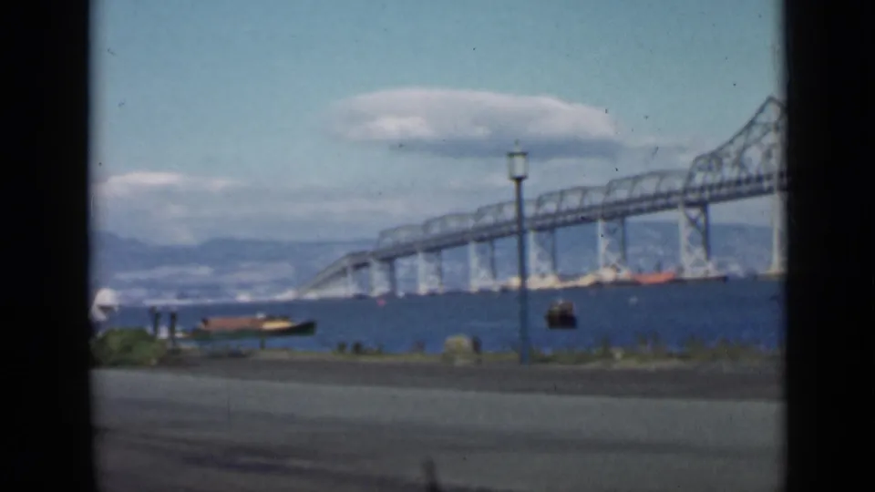 1939 San Francisco 8mm view of a bridge across the bay, with a seaplane near shore, a small boat, road, lamppost, distant hills, and clouds.