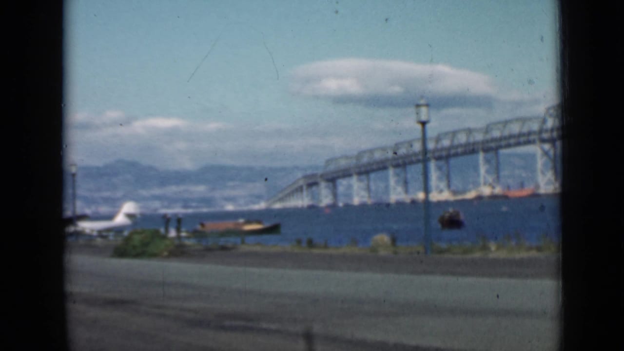 Bridge span over bay water with seaplane tail, small boat, lamppost, road, hills, and clouds.