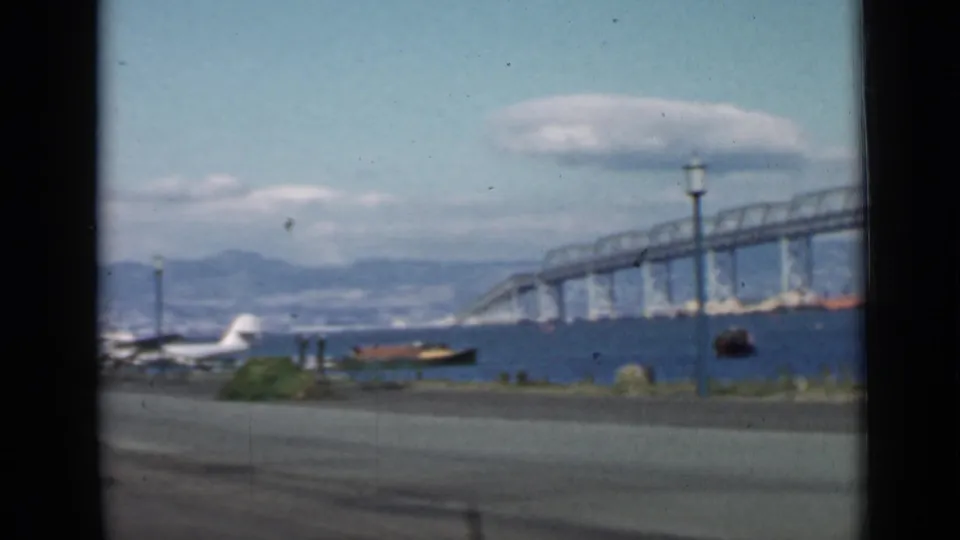 1939 San Francisco waterfront frame with a large bridge crossing the bay, a seaplane near shore, a small boat, road edge, lamppost, hills, and clouds.