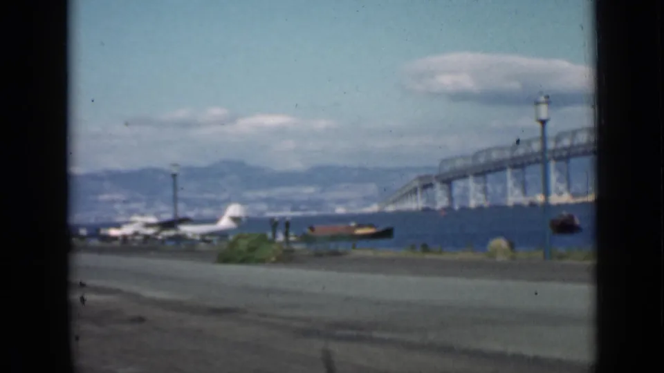 Final 1939 San Francisco 8mm frame showing the bridge over the bay, seaplane and boat near shore, road in front, lampposts, distant hills, and clouds.